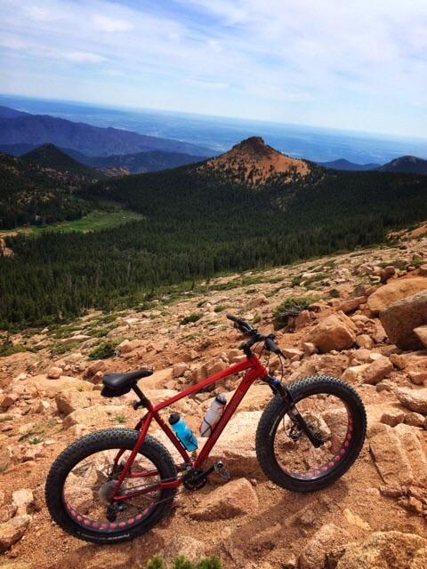 A red mountain bike with wide tires rests on rocky terrain overlooking a vast landscape of green forested hills and distant mountains under a cloudy sky. Elk Park Trail mountain bike trail.