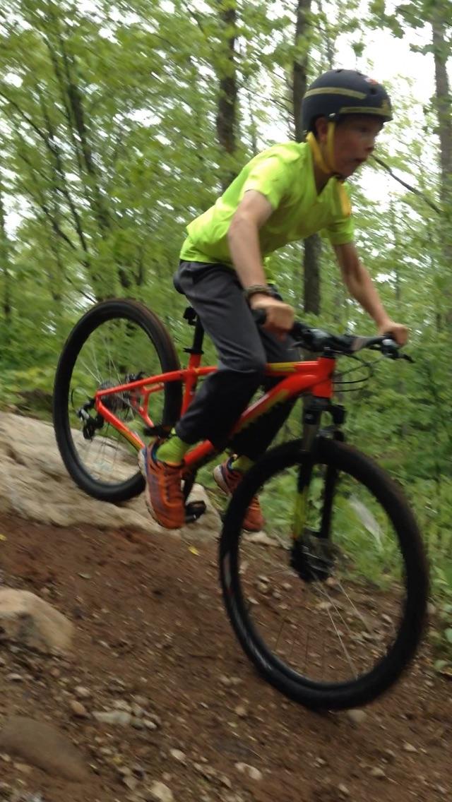 A young person riding a bright orange mountain bike on a dirt trail surrounded by lush green trees. The rider is airborne, jumping off a rocky surface, wearing a helmet and a fluorescent yellow shirt, with a focused expression. WinMan Trails mountain bike trail.