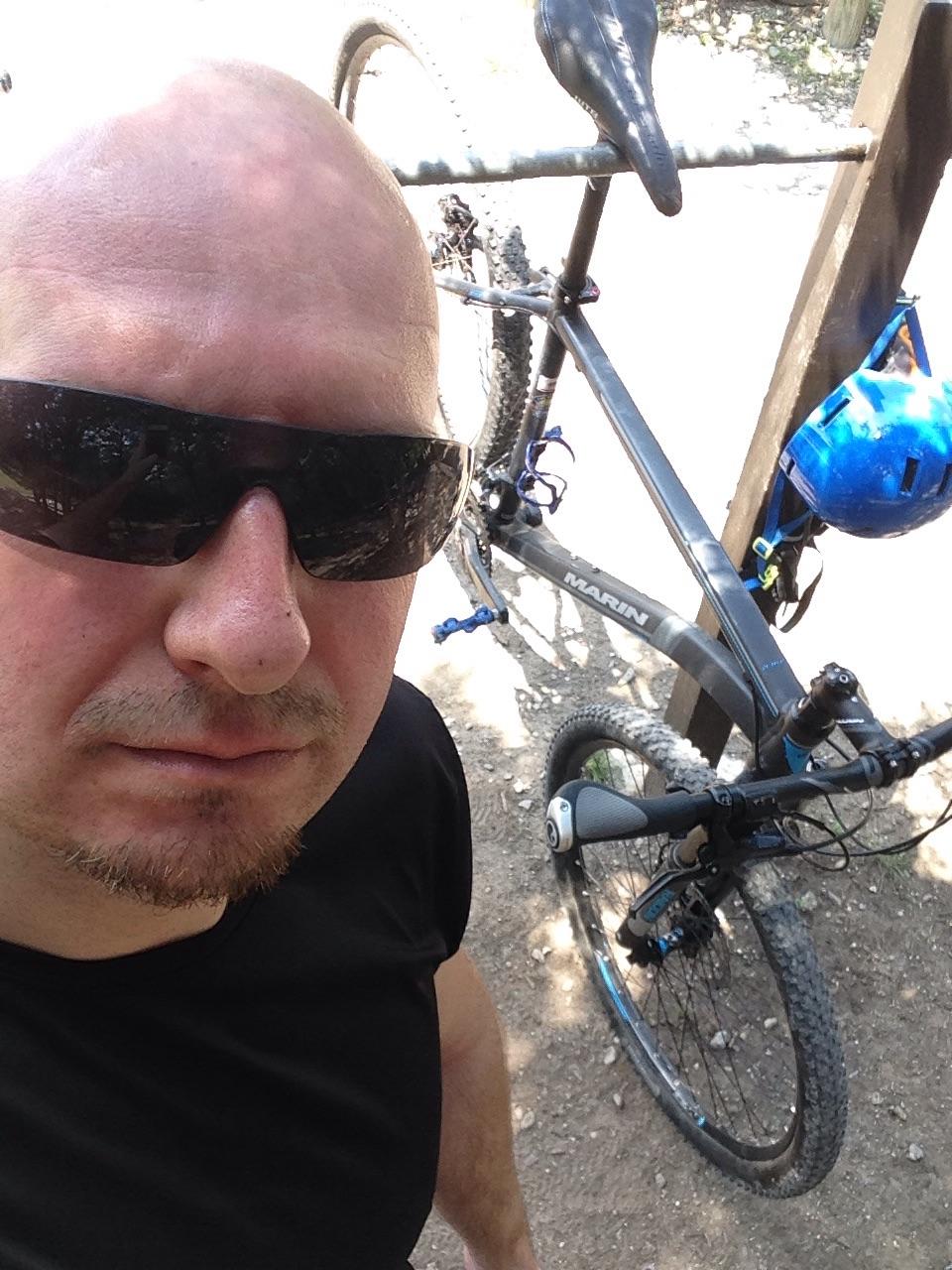 Marin Bobcat Trail: A close-up selfie of a man with a bald head, wearing dark sunglasses and a black shirt, standing next to a mountain bike. The background shows a dirt path and greenery, with the bike's blue helmet hanging from the handlebars. The image captures a sunny outdoor setting.