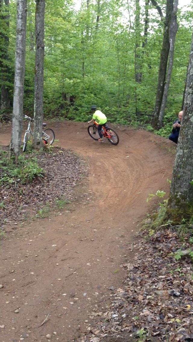 A child wearing a bright yellow shirt and a helmet is riding a bicycle around a dirt trail in a forested area. Two bicycles are positioned nearby, with lush green trees and foliage in the background. WinMan Trails mountain bike trail.