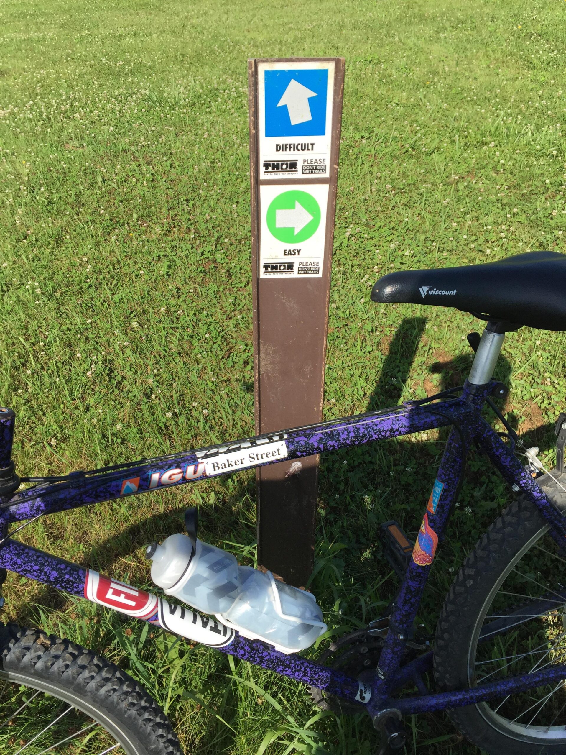 Giant Iguana: A mountain bike is parked next to a trail sign indicating difficulty levels for cyclists. The sign shows a blue arrow pointing up labeled "Difficult" and a green arrow pointing right labeled "Easy." The background features a grassy field.