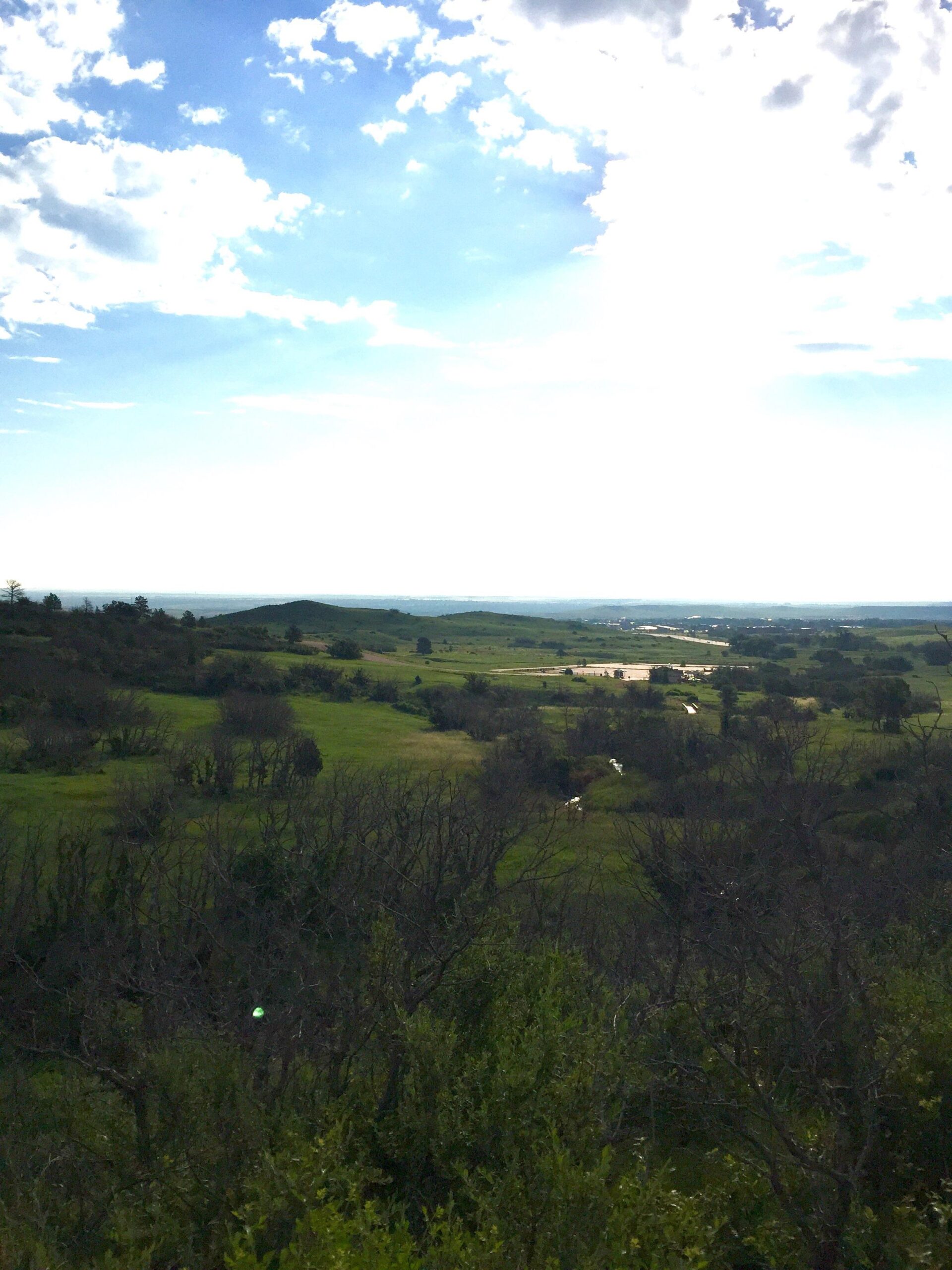 A panoramic view of a lush green landscape under a partly cloudy sky, featuring rolling hills and a winding river. The scene captures the tranquility of nature with patches of trees and a bright blue sky above. Cheyenne Mountain State Park mountain bike trail.