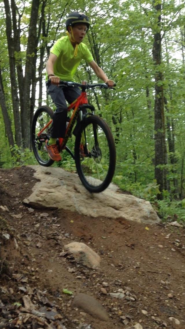 A young boy wearing a bright green shirt and a black helmet rides a mountain bike, airborne above a large rock on a forest trail. Trees and greenery fill the background, indicating an outdoor mountain biking scene filled with natural elements. WinMan Trails mountain bike trail.