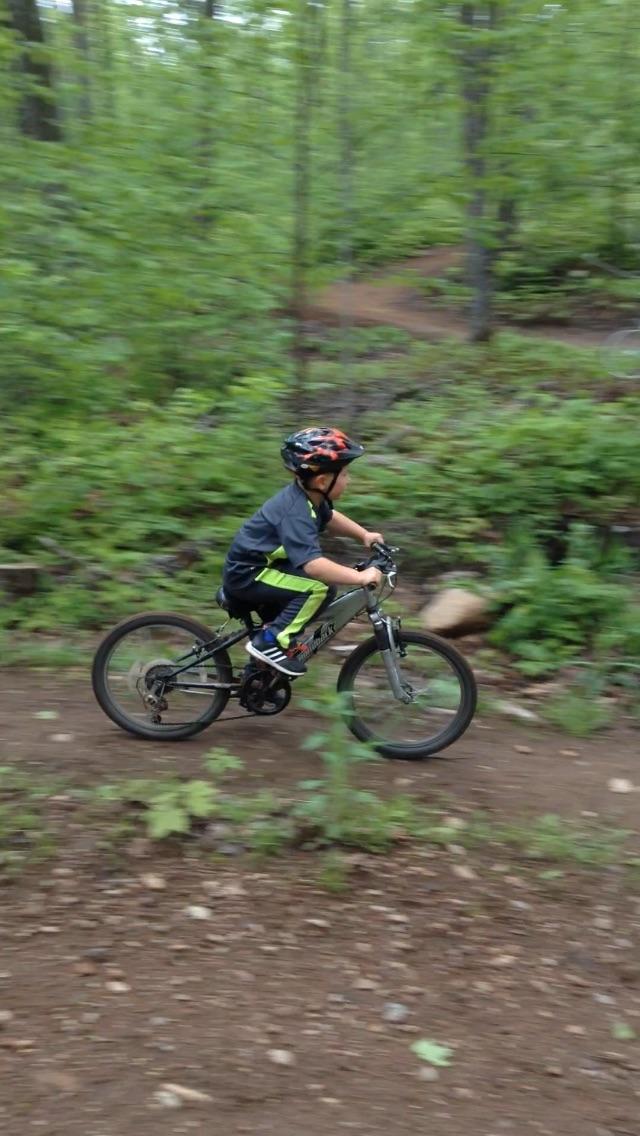 A child wearing a helmet rides a mountain bike along a dirt path in a lush green forest. The bike is in motion, showcasing speed as the surroundings blur slightly, highlighting the action. WinMan Trails mountain bike trail.