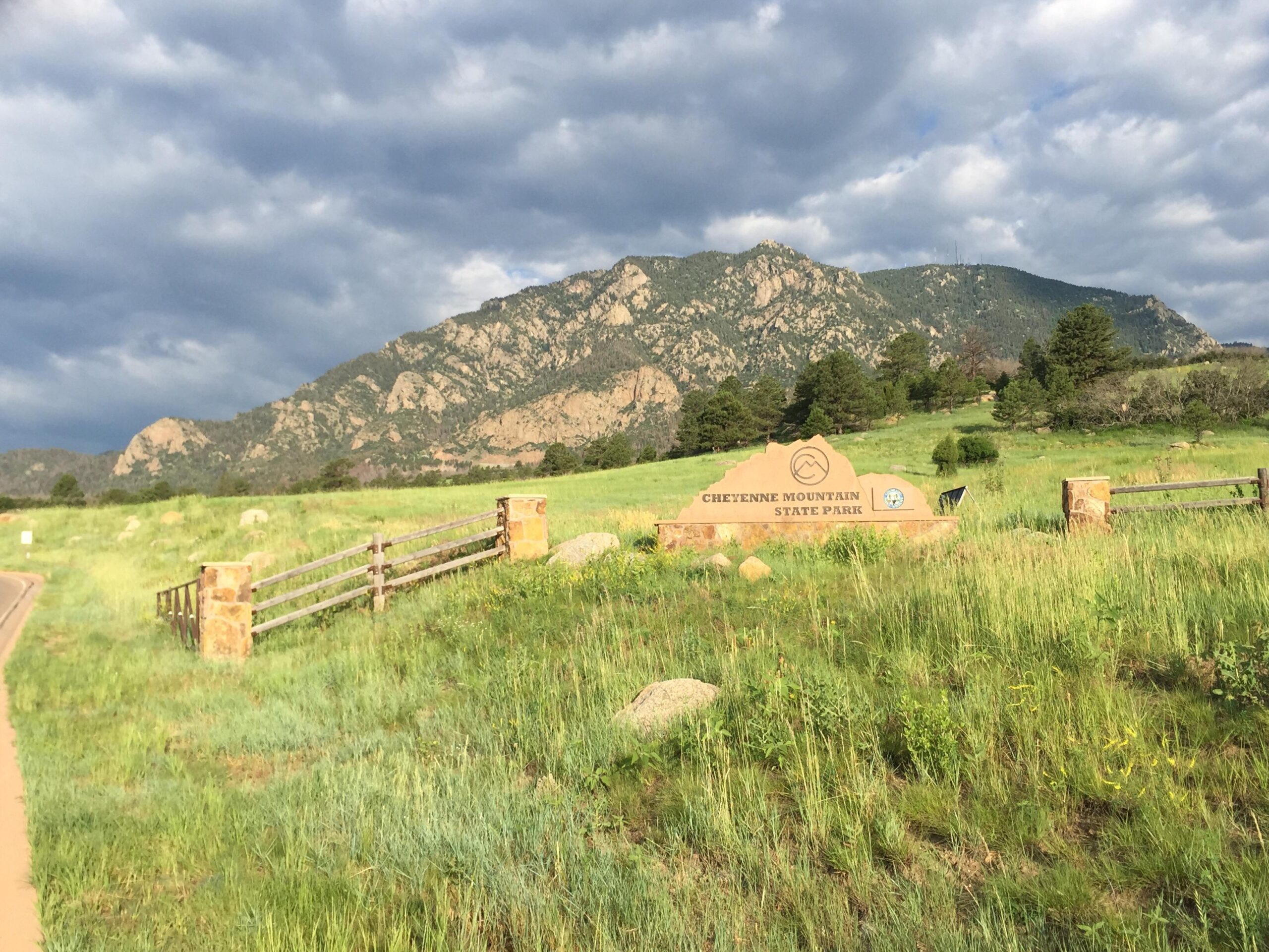 A scenic view of Cheyenne Mountain State Park entrance, featuring a stone sign with the park's name. The background showcases rugged mountains under a cloudy sky, with lush green grass and trees in the foreground. Cheyenne Mountain State Park mountain bike trail.