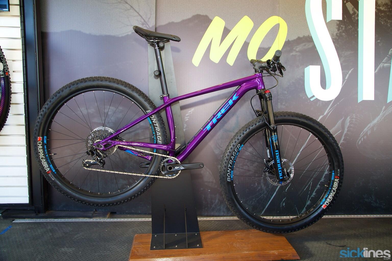 Trek Stache: A vibrant purple mountain bike displayed on a wooden stand against a backdrop featuring the words "MO ST" and a mountain graphic. The bike has wide tires, a sleek frame, and components branded with Bontrager and Manitou logos.