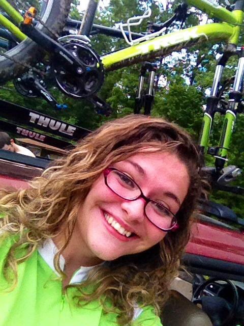 A person with curly hair, wearing glasses and a bright green shirt, smiles for a selfie in front of a vehicle with two bicycles mounted on a roof rack. Lush green trees are visible in the background, indicating an outdoor setting. Carvin's Cove Trail system mountain bike trail.
