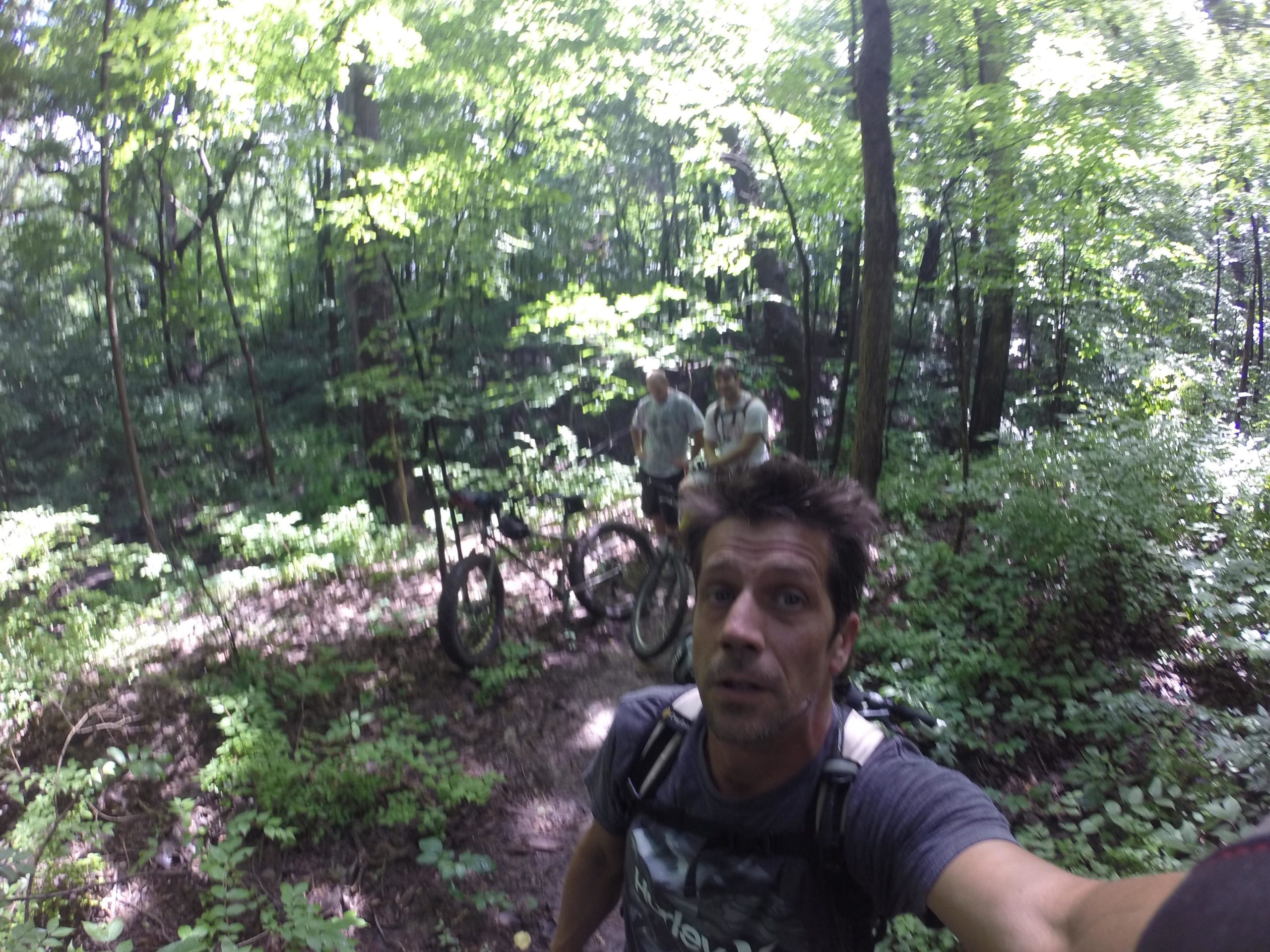 A group of three individuals in a lush green forest, with two people standing near their mountain bicycles in the background. The foreground features a person taking a selfie, surrounded by dense foliage and sunlight filtering through the trees. Murdock Park Trail mountain bike trail.