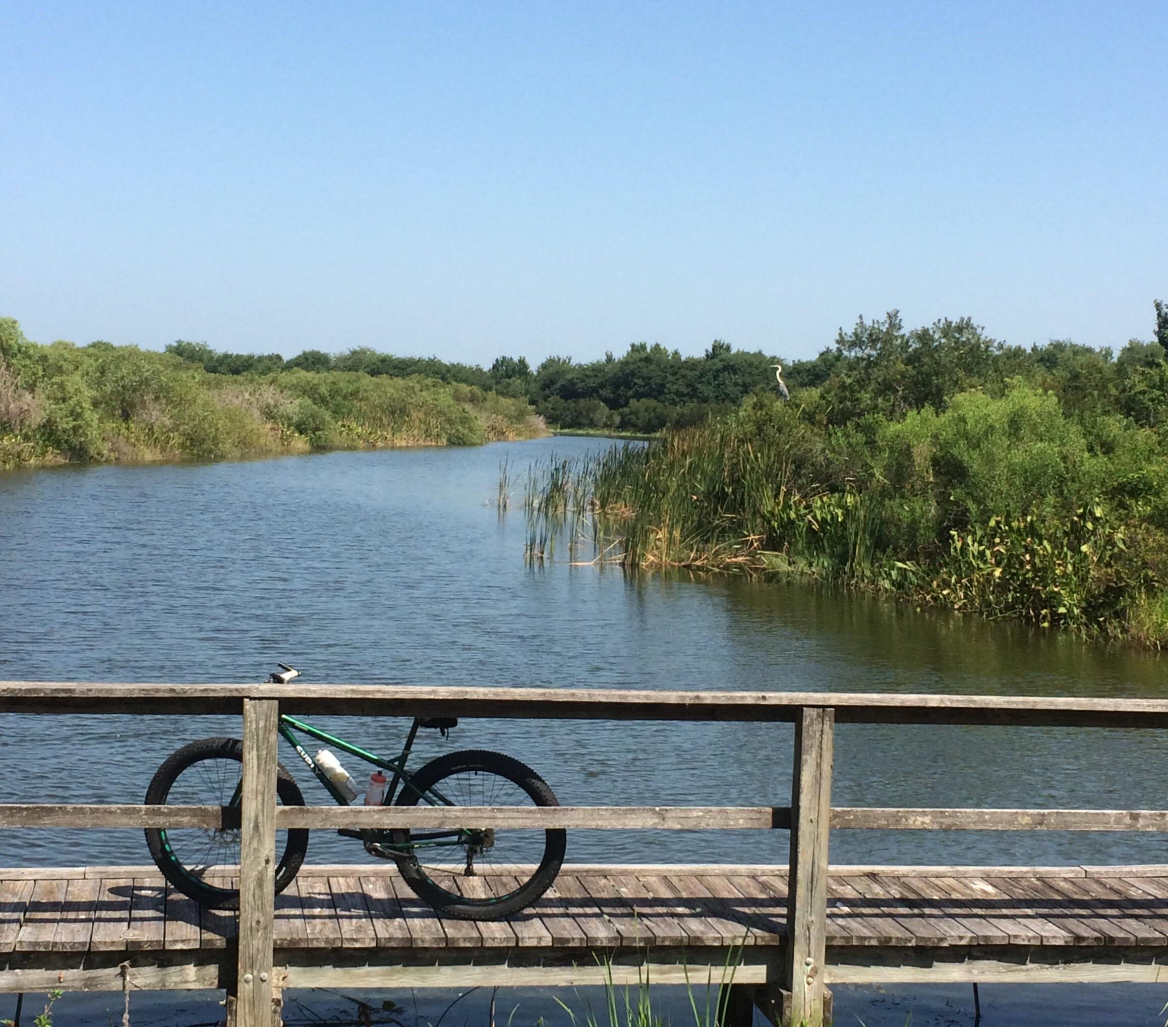 A mountain bike resting on a wooden bridge over a calm river, surrounded by lush greenery and tall grasses under a clear blue sky. Lake Apopka Restoration Area mountain bike trail.