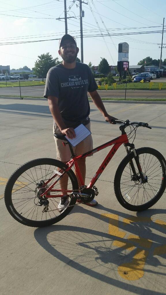 Trek Marlin 5: A smiling man standing next to a red mountain bike in a parking lot, wearing a gray shirt and beige shorts, holding a piece of paper in his left hand. The background features greenery, power lines, and parked cars under a clear sky.