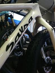 Salsa Beargrease: Close-up of a white bicycle frame featuring the brand name "Sauza," with a focus on the front fork and wheel. The background shows additional bicycles and equipment in a shop setting.