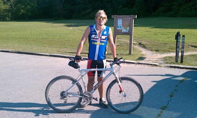 GT Zaskar: A person wearing a blue cycling jersey and sunglasses stands beside a silver mountain bike on a paved path. In the background, there is a grassy area with a wooden information board and bike trail markers, indicating a recreation or biking area.