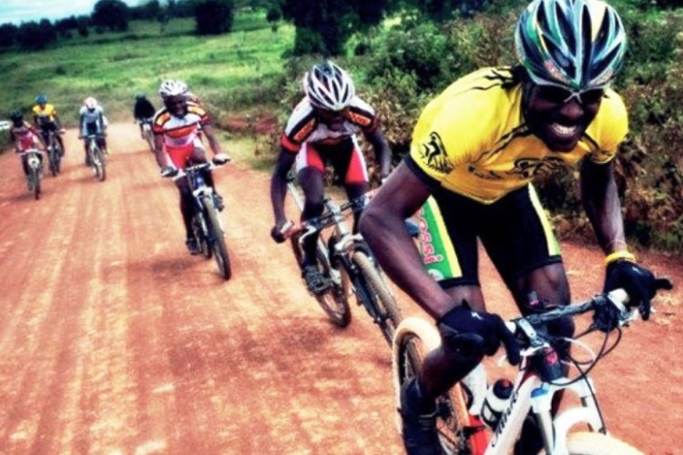 A group of six cyclists riding on a red dirt road surrounded by green fields. The cyclists are wearing colorful jerseys and helmets, showcasing a mix of athleticism and enthusiasm as they navigate the terrain. The scene captures the excitement of mountain biking in a natural setting.