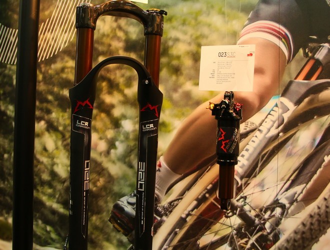Image of a mountain bike suspension fork and a shock, displayed against a backdrop featuring a cyclist. The fork has a black design with red and silver branding, and the shock is smaller, with similar color accents. A product information card is visible, providing details about the items.