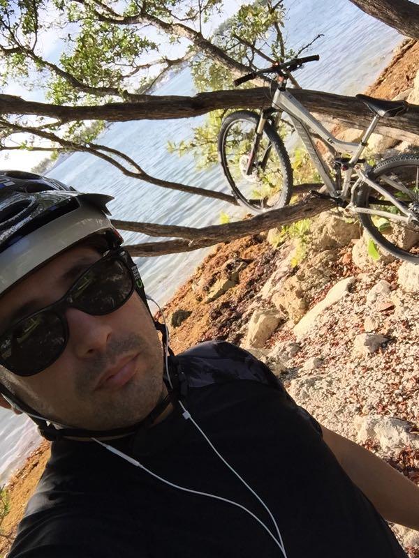 A person wearing a helmet and sunglasses stands near the water's edge, with earbuds in their ears. In the background, a mountain bike is perched on a tree branch, surrounded by rocks and vegetation. The scene features a calm body of water and a sunny sky. Oleta River State Park mountain bike trail.