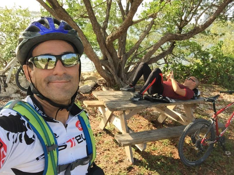 A man wearing a helmet and reflective sunglasses smiles for a selfie while posing next to a picnic table in a natural setting. Another man relaxes on the table, using a mobile device. Bicycles are parked nearby, and trees and a body of water are visible in the background. Oleta River State Park mountain bike trail.