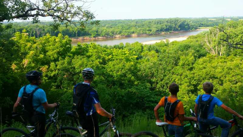 Four mountain bikers pause to enjoy a scenic view of a river surrounded by lush greenery. They stand on a hillside, looking out over the landscape, with trees in the foreground and the river winding through the background under a clear sky. Camp Horizon mountain bike trail.