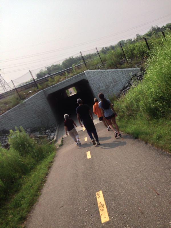A group of five individuals walking along a paved path that leads to a tunnel. The path is surrounded by tall grass and greenery, with power lines visible in the background. The sky appears hazy, indicating a warm day. Kal-Haven Trail mountain bike trail.