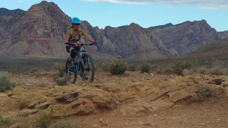 A mountain biker in a blue helmet navigates rocky terrain with rugged mountains in the background, showcasing an outdoor adventure setting. Cottonwood Valley North mountain bike trail.