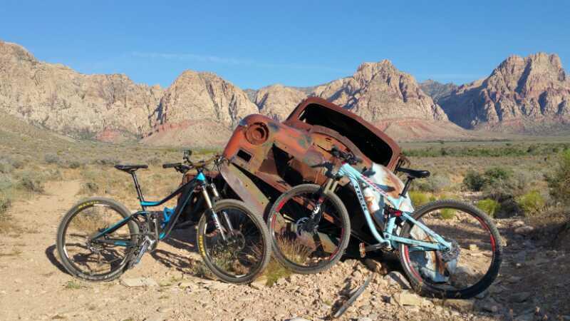 Two mountain bikes leaning against a rusted, abandoned vehicle in a desert landscape, with rugged mountains in the background under a clear blue sky. Blue Diamond mountain bike trail.