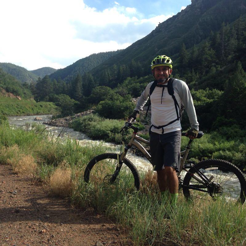 A person wearing a green helmet and cycling gear stands next to a mountain bike on a dirt path by a flowing river. Lush green vegetation and mountains are in the background, under a partly cloudy sky. Waterton Canyon mountain bike trail.