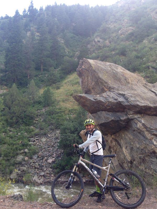 A person in biking gear standing next to a mountain bike beside a rocky landscape with trees and a stream in the background. The rider is wearing a helmet and smiling, with a large rock formation nearby. Waterton Canyon mountain bike trail.