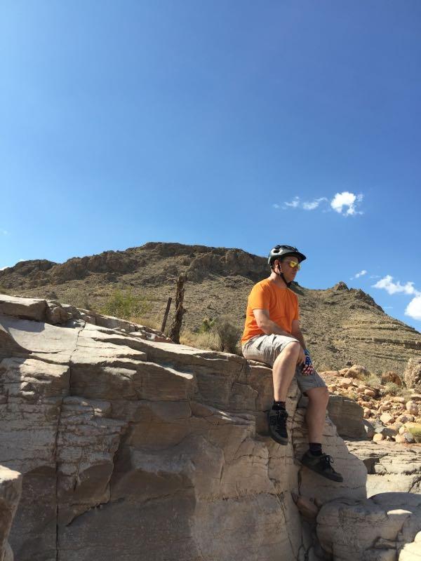 A person in an orange shirt and black helmet sits on a rock ledge, overlooking a desert landscape with mountains in the background and a clear blue sky above. The scene depicts outdoor adventure and exploration. Blue Diamond mountain bike trail.