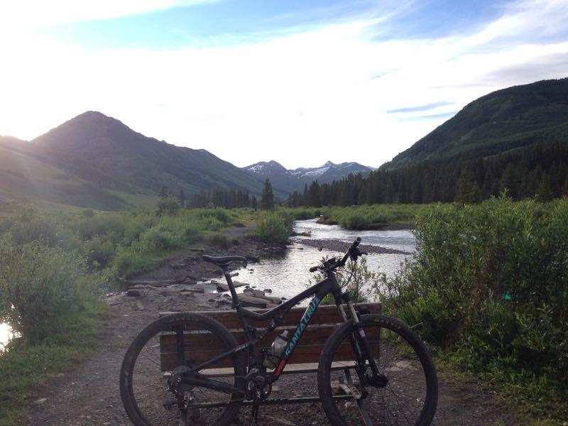 Mountain bike resting on a wooden bench near a river, with lush greenery, mountains, and a partially cloudy sky in the background. Lower Loop mountain bike trail.