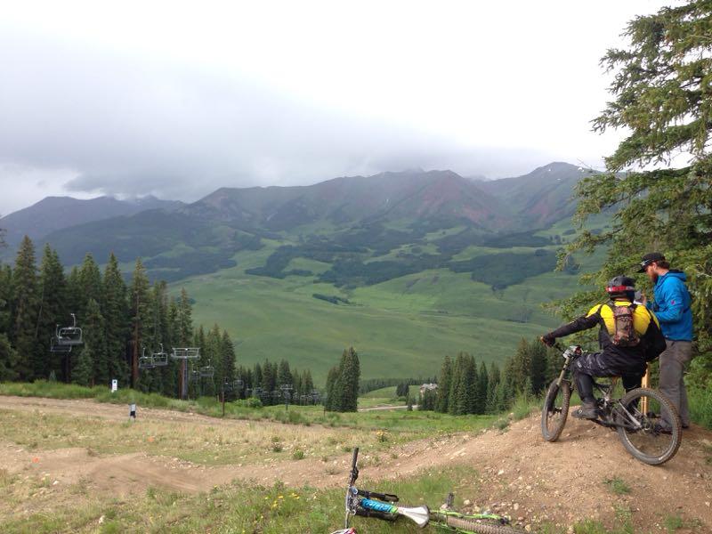 A scenic view from a mountain biking trail showing two cyclists overlooking a lush valley surrounded by mountains. In the foreground, one cyclist is seated on a bike while the other stands next to him. Several pine trees and a ski lift can be seen in the background, under a cloudy sky. Wood's Trail mountain bike trail.