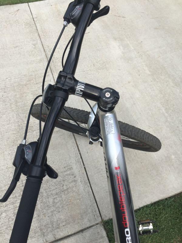 A close-up view of a mountain bike's handlebars and front fork, showing the grips, brake levers, and shifters. The image captures the bike resting on a paved surface with grass partially visible in the background. Augusta Canal mountain bike trail.