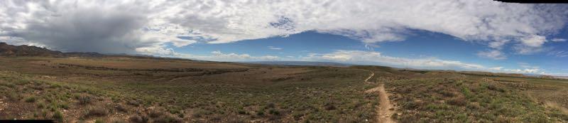 Panoramic view of a grassy landscape under a partly cloudy sky, featuring rolling hills and a dirt path winding through the terrain. Western Zippety mountain bike trail.
