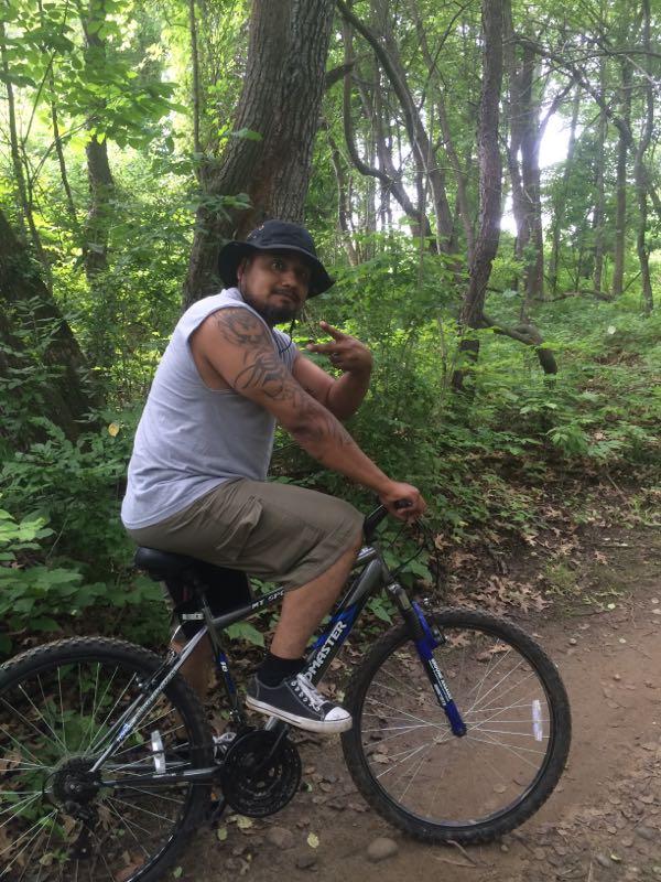 A man with tattoos is sitting on a mountain bike on a dirt trail in a lush green forest. He is wearing a gray sleeveless shirt and tan shorts, and he is giving a peace sign with his right hand while smiling at the camera. Addison Oaks mountain bike trail.