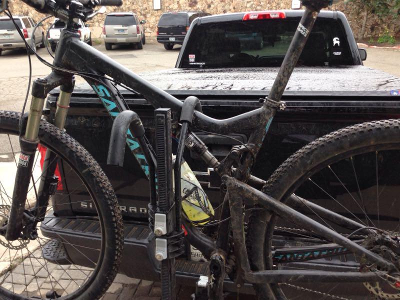 A close-up view of a muddy mountain bike secured to the back of a black pickup truck. The bike features a suspension system, visible dirt on the frame and tires, and a colorful water bottle attached to the frame, with a blurred background of parked vehicles. Trail 401 mountain bike trail.