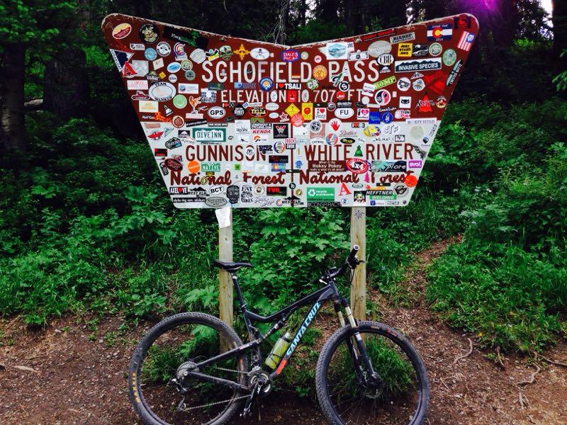 A wooden sign at Schofield Pass, adorned with numerous colorful stickers, indicating its elevation of 10,707 feet and the surrounding Gunnison and White River National Forests. In front of the sign, a mountain bike is parked on the trail, surrounded by lush green foliage. Trail 401 mountain bike trail.