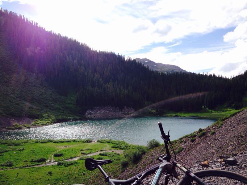 A scenic view of a tranquil lake surrounded by lush green hills and dense trees, with a bicycle resting in the foreground. The sunlight reflects off the water, creating a sparkling effect. Trail 401 mountain bike trail.