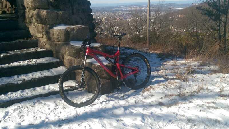 A red mountain bike leaning against a stone structure on a snowy hillside, with a distant view of a city in the background. Green Lane mountain bike trail.