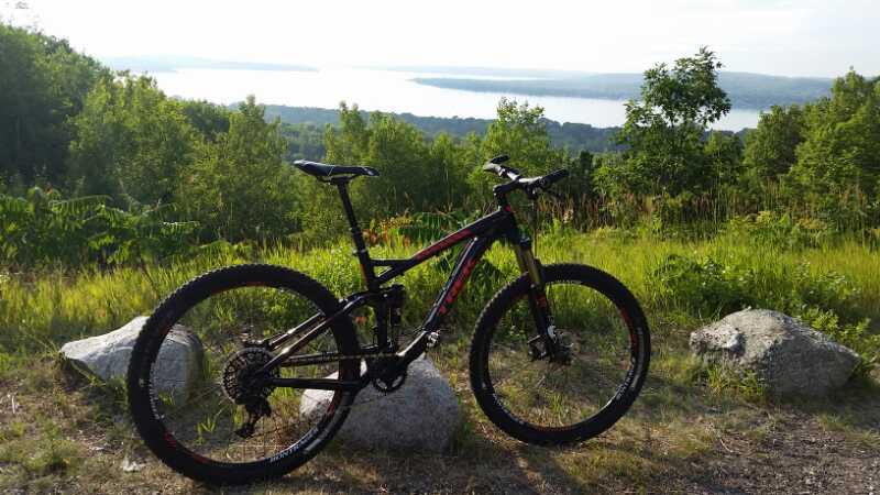 Mountain bike resting on rocks with a scenic view of a lake and dense greenery in the background during daytime. Avalanche Preserve mountain bike trail.