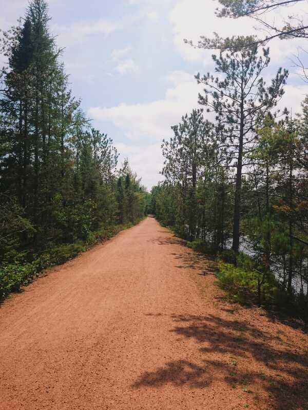 A dirt path surrounded by tall trees on both sides, leading toward a tranquil body of water. The sky is bright with a few clouds, creating a serene outdoor atmosphere. Bearskin State Trail mountain bike trail.
