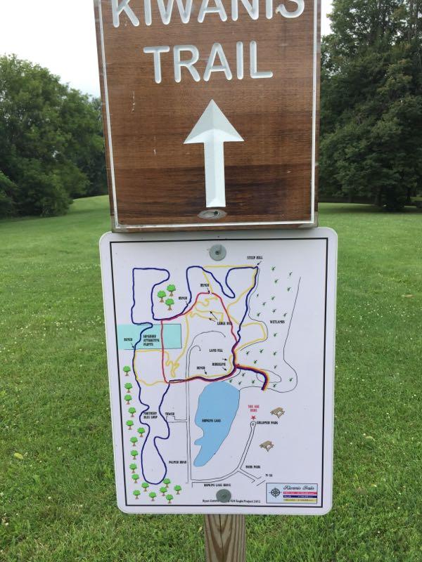 Sign for Kiwanis Trail with an arrow pointing upward, accompanied by a map detailing the trail routes. The map features paths highlighted in different colors, surrounding areas such as ponds and shaded regions, with labeled landmarks and symbols for trees and wildlife. The setting is a grassy area with trees in the background. kiwanis trail mountain bike trail.