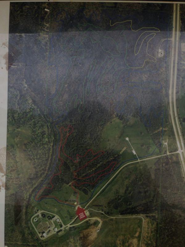 A topographic map depicting a park layout with colored trail markings. The trails are outlined in red, blue, and green, indicating different routes through a forested area. A labeled area for parking and facilities is located at the bottom, near a prominent red marker indicating "You Are Here." The surrounding landscape features varying terrain with patches of greenery and open fields. An adjacent road is visible on the right side of the image. Sac River Trail mountain bike trail.