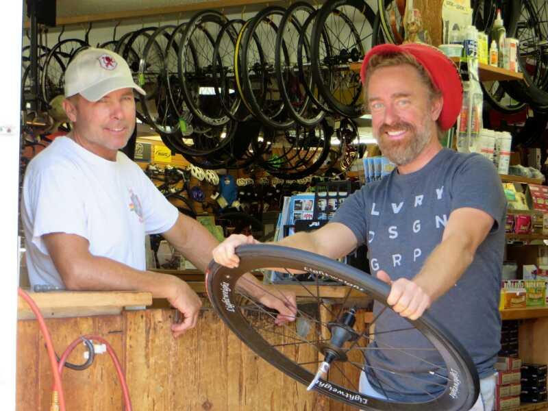 Two men are standing inside a bicycle shop. One man, wearing a white t-shirt and a cap, is smiling and leaning against the counter. The other man, wearing a blue t-shirt and a red hat, is holding a bicycle wheel and grinning. In the background, various bicycle wheels and accessories can be seen displayed on the shop walls.