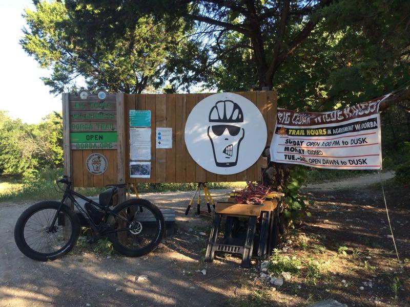 Wooden signboard at a trail entrance featuring a large graphic of a skull wearing sunglasses and a bike helmet. Nearby, a black bicycle is leaning against the sign. Information about trail hours and rules is posted on the signboard, and there is a banner indicating that the trails are open and providing specific hours of operation. The setting is surrounded by trees, suggesting a natural environment. Big Cedar Wilderness Trails mountain bike trail.