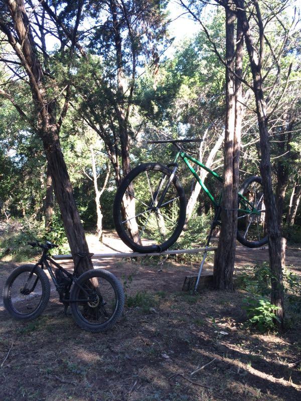 A green mountain bike is suspended between two trees, with one wheel resting on a horizontal bar. A black mountain bike is positioned on the ground nearby, surrounded by a wooded area with sunlight filtering through the trees. Big Cedar Wilderness Trails mountain bike trail.