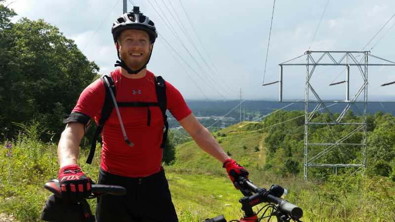 A smiling cyclist wearing a red athletic shirt and black gloves is standing next to a mountain bike on a grassy hill. In the background, there are power lines and a view of a valley surrounded by trees under a cloudy sky. Coldwater Mountain mountain bike trail.