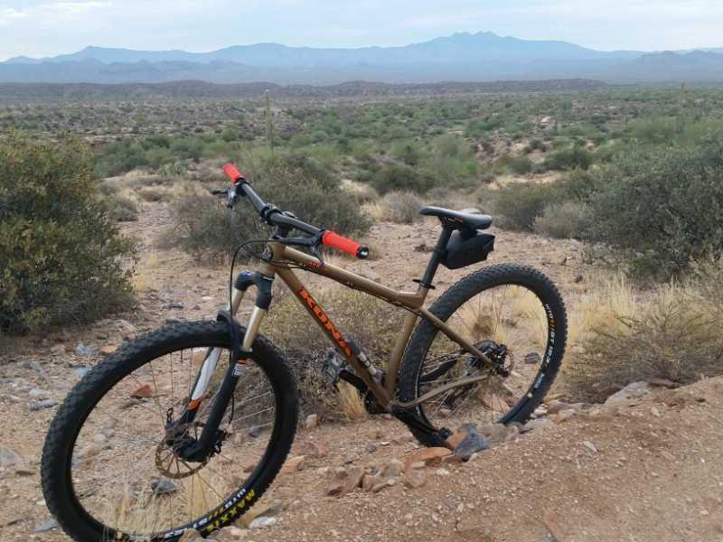 A mountain bike rests on a rocky trail, surrounded by desert vegetation and rolling hills under a cloudy sky. The background features distant mountains, while the bike has a mix of dark and brown tones with red grips. McDowell Mountain Park mountain bike trail.