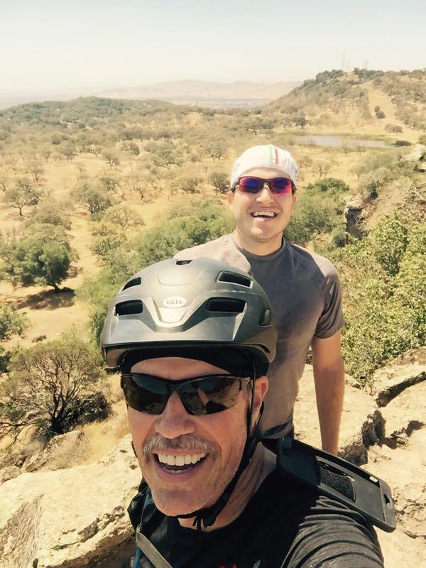 Two men smiling for a selfie while biking outdoors. One man is wearing a black helmet and sunglasses, and the other is in a gray shirt with a headband. They are standing on a rocky outcrop with a scenic view of a hilly landscape filled with trees and a distant lake. The sun is shining brightly overhead, suggesting a warm day. Rockville Park mountain bike trail.