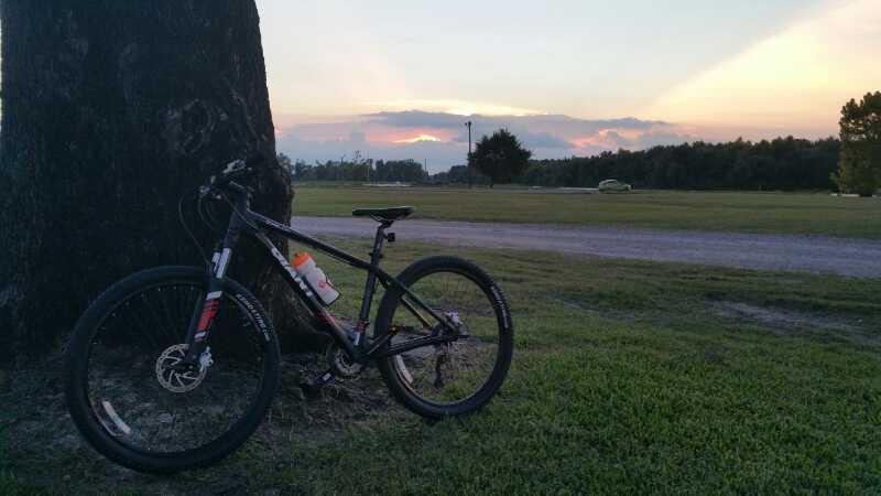 A mountain bike leaning against a large tree at sunset, with a grassy field and a pathway in the background. The sky is filled with clouds and soft colors, creating a peaceful outdoor atmosphere. Bonnet Carre Spillway Trail mountain bike trail.