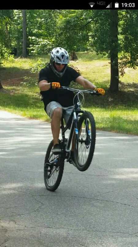 A person riding a mountain bike performs a wheelie on a paved road surrounded by green trees. They are wearing a helmet and gloves, with a determined expression on their face as they maintain balance on the back wheel of the bike. Flat Rock Park mountain bike trail.