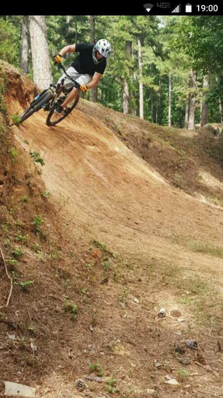 A mountain biker skillfully navigating a dirt trail with a downward slope. The rider, wearing a helmet and protective gear, leans into the turn, showcasing dynamic movement. Lush greenery and trees are visible in the background, creating a natural setting. Pine cones and smaller plants can be seen on the ground near the trail. Flat Rock Park mountain bike trail.