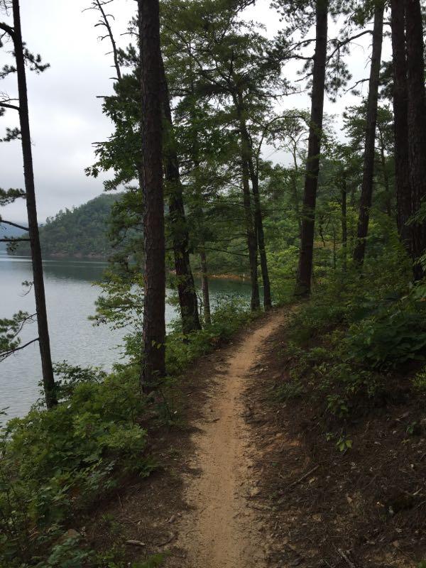 A winding dirt path bordered by trees leads towards a calm lake, surrounded by mountains under a cloudy sky. Tsali Recreation Area mountain bike trail.
